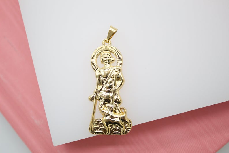 A detailed gold pendant displays Saint Roch holding a staff and standing beside a faithful dog against a white surface.