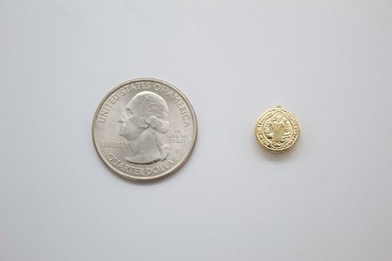 A small gold Saint Benedict medal rests beside a United States quarter coin on a light surface to show its miniature size.