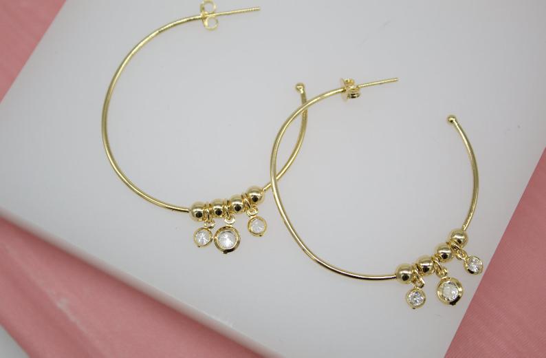 A pair of gold hoop earrings features dangling cubic zirconia charms on a white display surface.
