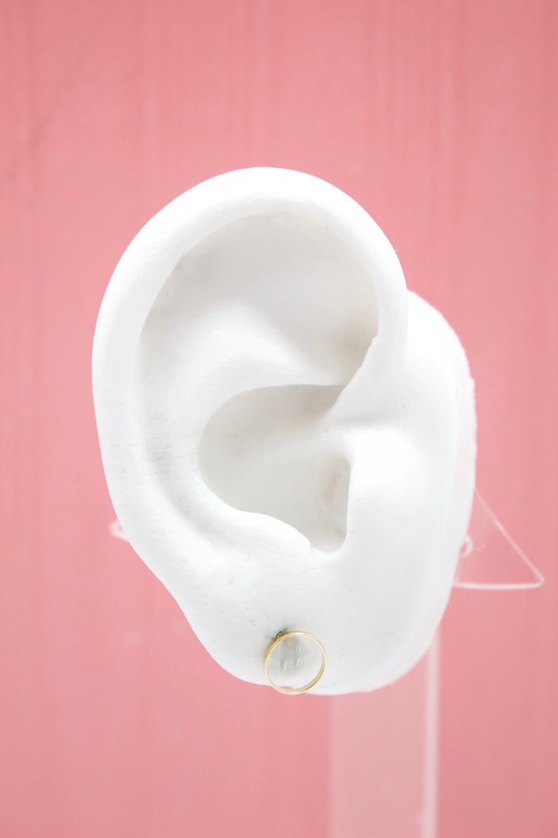 A white mannequin ear displays a minimalist gold hoop earring against a pink background.