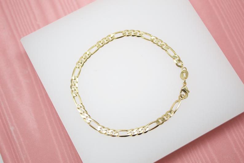 A delicate gold Figaro link chain bracelet rests on a white display surface against a pink background.
