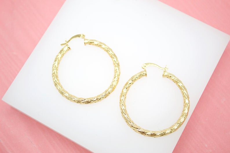 A pair of textured gold hoop earrings rests neatly on a white display surface against a soft pink background.