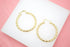 A pair of textured gold hoop earrings rests neatly on a white display surface against a soft pink background.