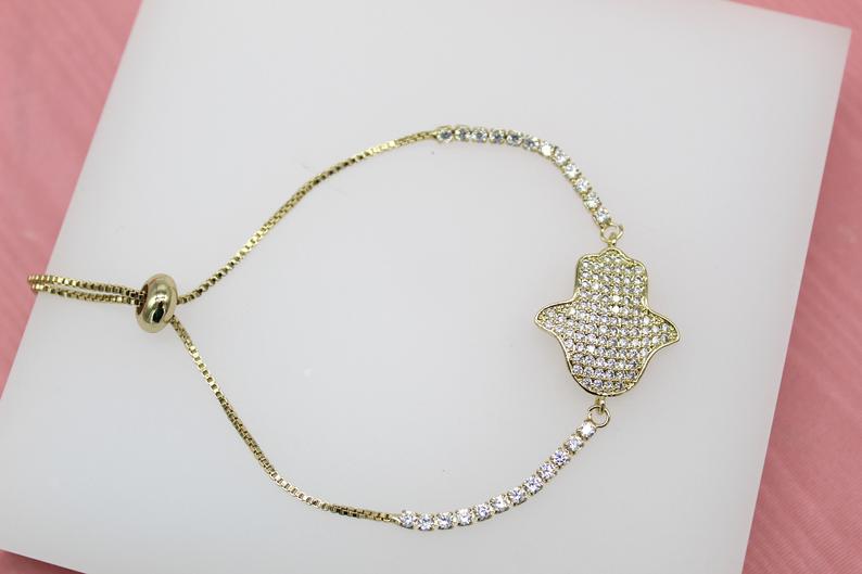 A gold Hamsa hand slider bracelet embellished with sparkling cubic zirconia crystals rests on a white display surface.