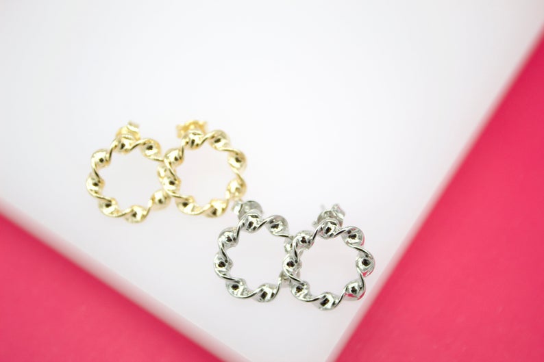 Two pairs of twisted hoop earrings, one gold and one silver, rest on a white surface against a bright pink background.