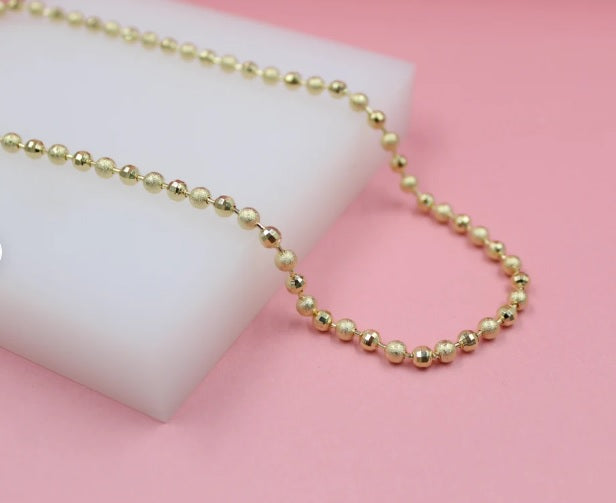 A delicate gold ball chain necklace rests gracefully on a white display block against a soft pink background.
