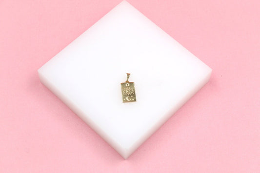 A gold rectangular pendant featuring celestial engravings of an eye, sun, and moon rests on a white display block against a soft pink background.