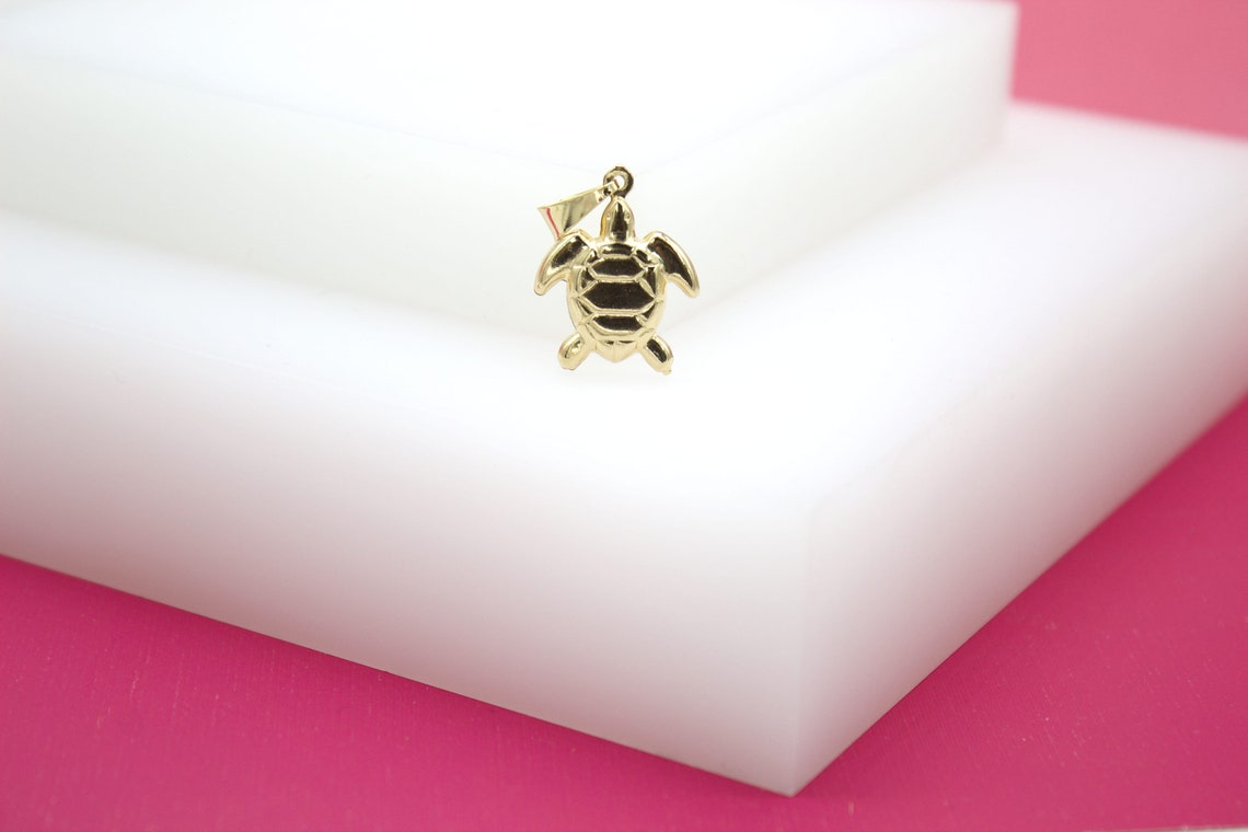 A gold turtle pendant rests on a white display block against a pink background.