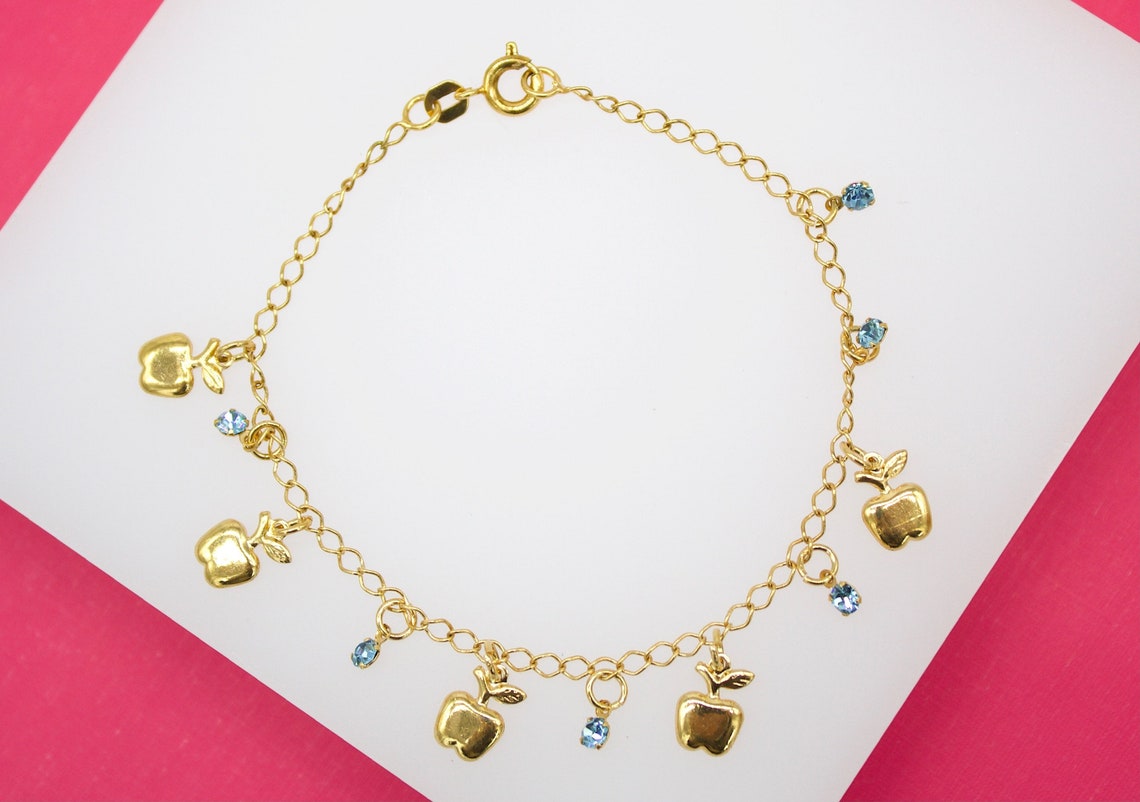 A delicate gold charm bracelet displays hanging apple charms interspersed with sparkling blue crystal gemstones against a white and pink background.