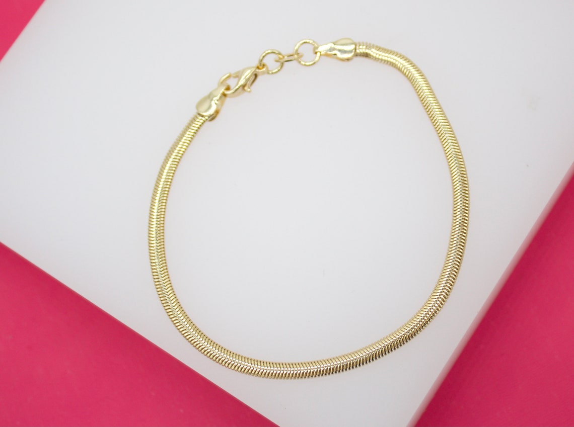 A delicate gold snake chain bracelet rests in a circle on a white display surface against a bright pink background.