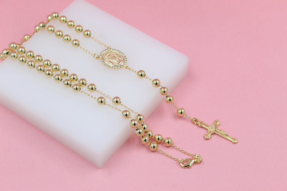 A polished gold rosary necklace rests gracefully on a white display block against a soft pink background.