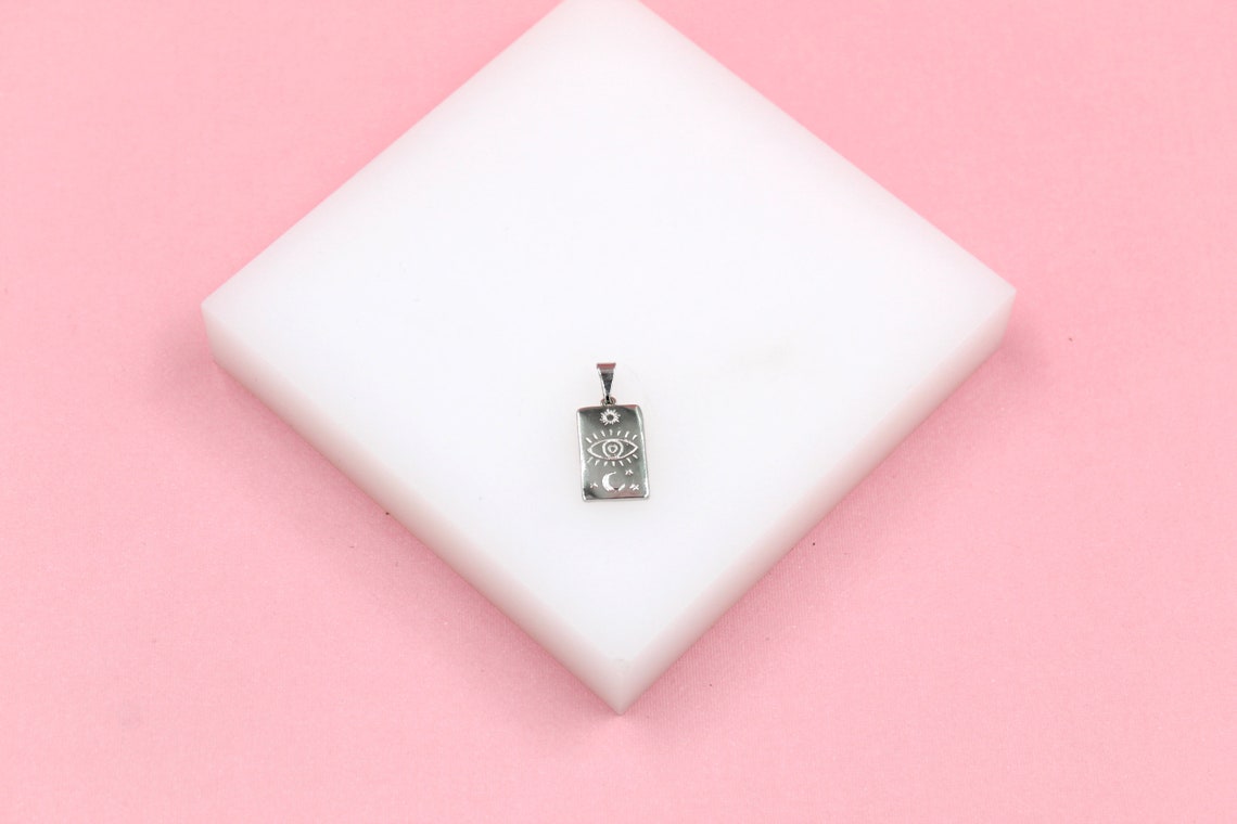 A silver rectangular pendant featuring a mystical evil eye, sun, and moon design rests on a white display block against a soft pink background.