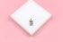 A rectangular silver pendant featuring an engraved evil eye, sun, and moon design rests on a white square display against a pink background.