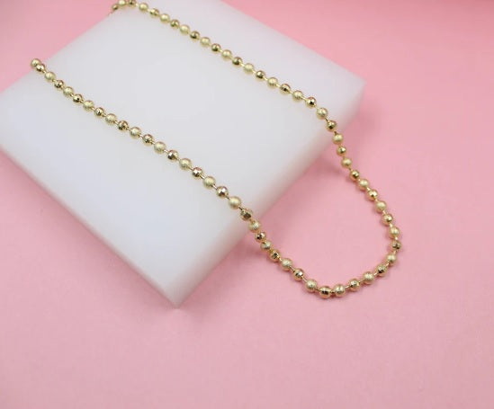 A delicate gold ball chain necklace rests on a white display block against a soft pink background.