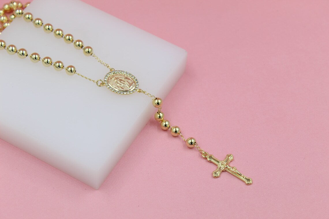 A gold rosary necklace with polished beads and a crucifix rests on a white display block against a pink background.