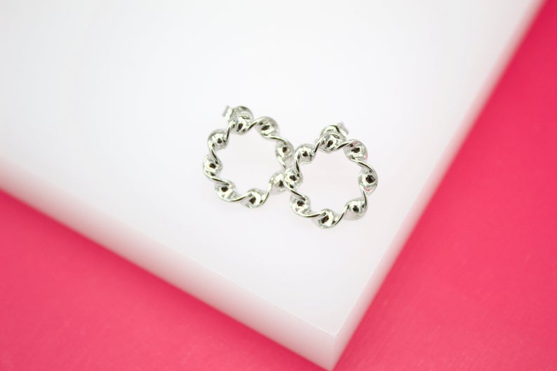 A pair of shiny silver twist hoop earrings rests on a white display surface against a vibrant pink background.