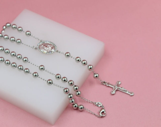 A gleaming silver rosary necklace with polished bead chains and a detailed crucifix rests on a white display block against a soft pink background.