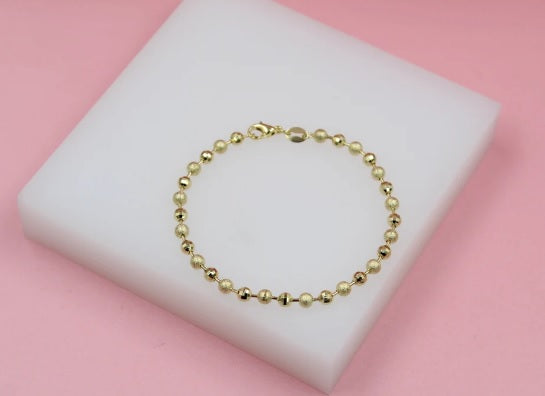 A delicate gold ball chain bracelet rests on a white display block against a soft pink background.