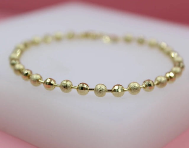 A delicate gold bead bracelet lies curved on a white display surface against a soft pink background.