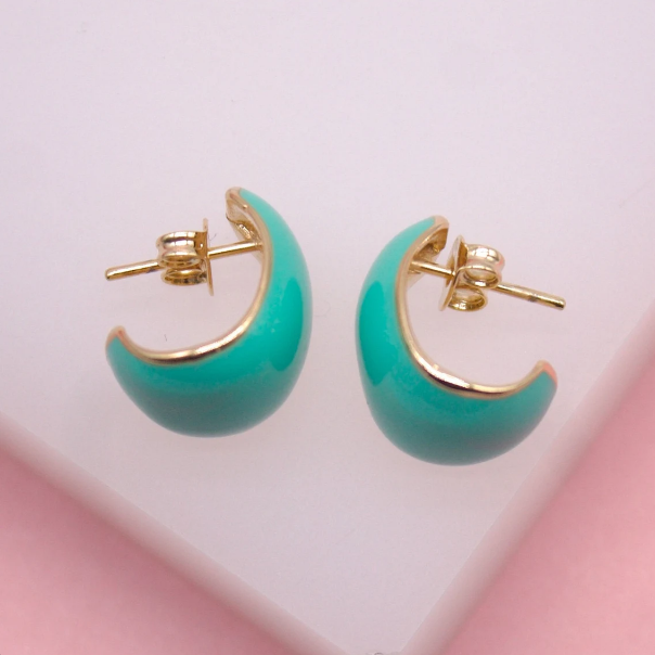 A pair of vibrant turquoise enameled hoop earrings rests on a light pink surface, showcasing their gold trim and secure post backs.