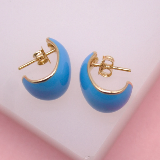 A pair of vibrant blue enamel hoop earrings with gold backing rests on a soft pink surface.