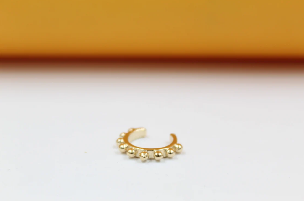 A shiny gold-plated ear cuff rests on a white surface, showcasing a beaded texture along its curved edge.