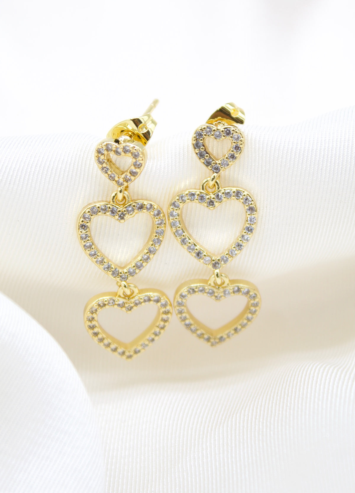 A pair of gold triple heart drop earrings encrusted with sparkling cubic zirconia stones rests elegantly on white satin fabric.