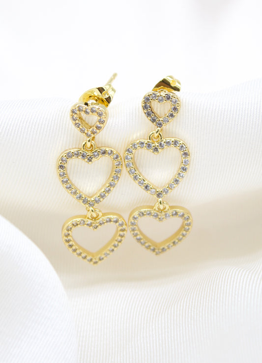 A pair of gold triple heart drop earrings encrusted with sparkling cubic zirconia stones rests elegantly on white satin fabric.