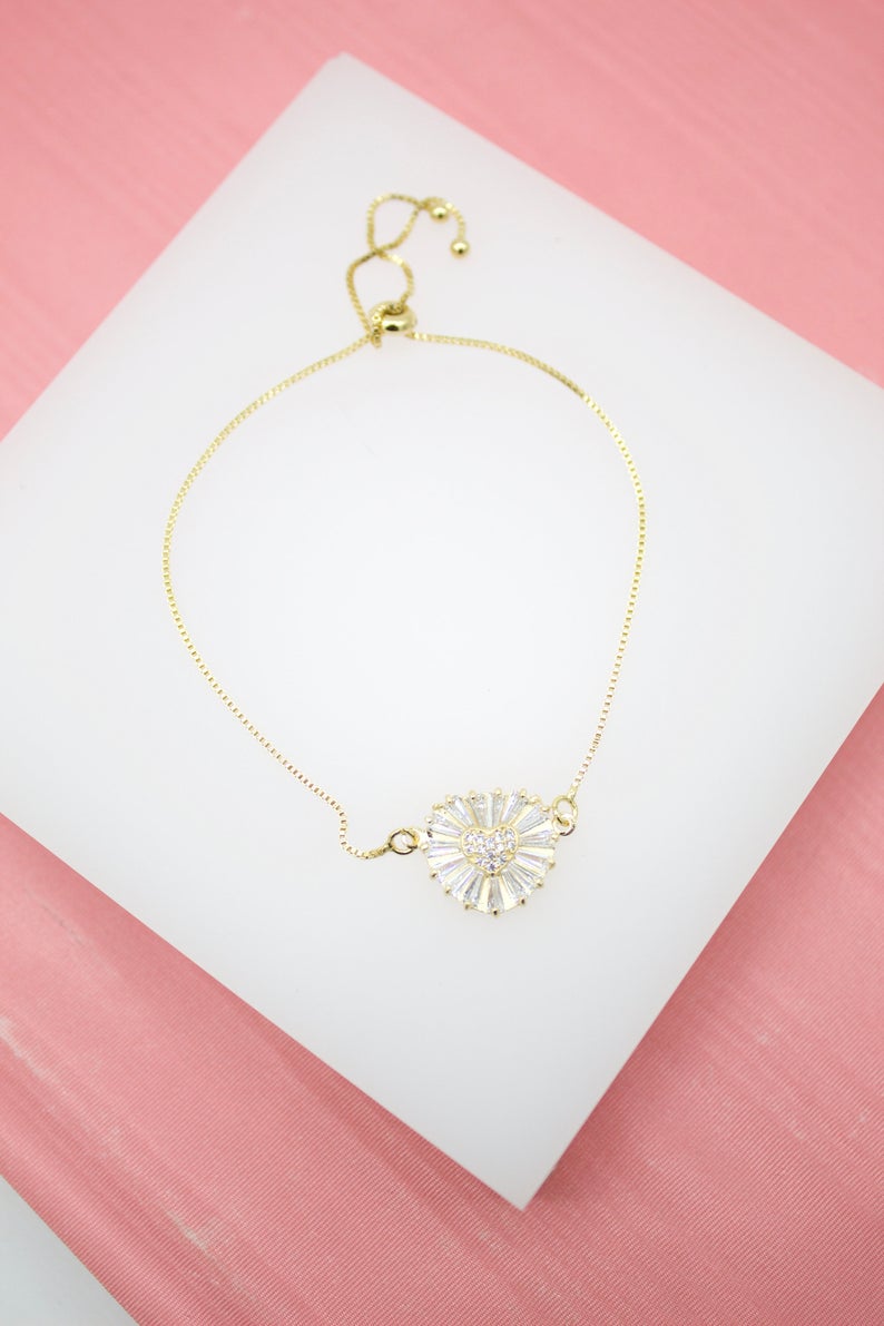 A delicate gold adjustable bracelet features a sparkling crystal heart pendant centered on a white display surface against a pink background.