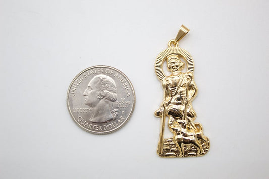 A gold Saint Lazarus pendant with a dog stands next to a US quarter coin for scale on a light gray surface.