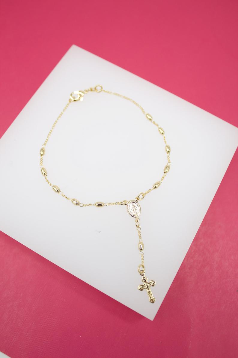 A delicate gold rosary bracelet rests on a white display surface against a vibrant pink background.
