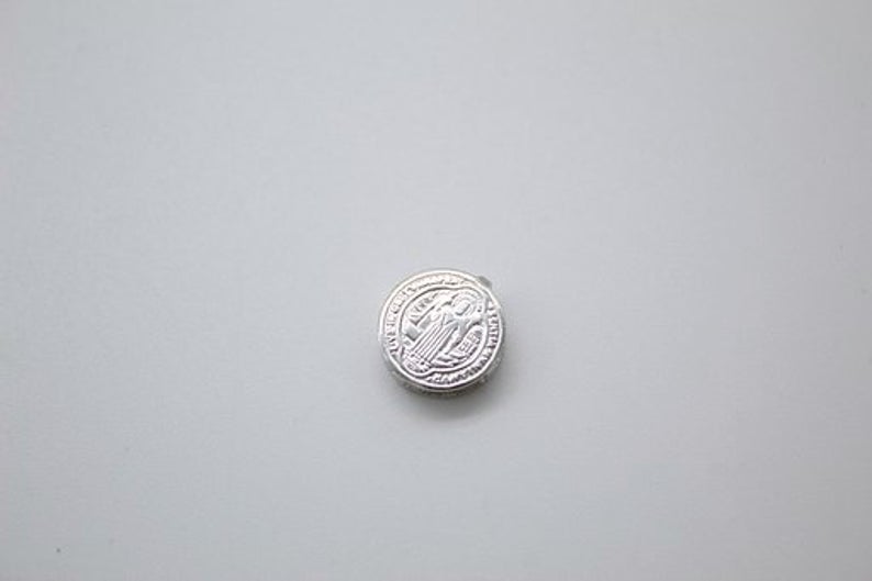 A small silver Saint Benedict medal rests centered on a neutral gray background.