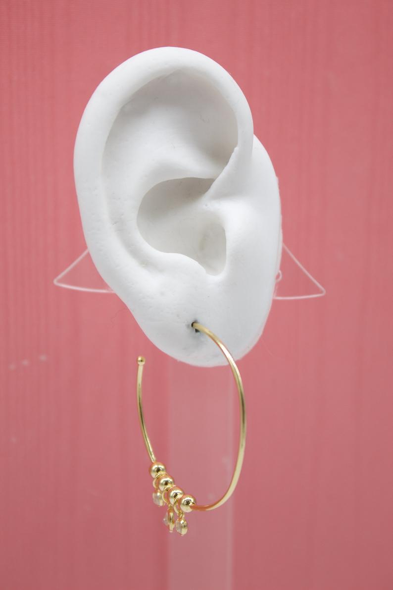 A white mannequin ear displays a gold beaded hoop earring against a pink background.
