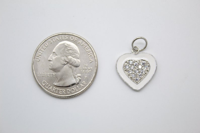 A white enamel heart charm with a pavé crystal center rests next to a US quarter for size comparison.