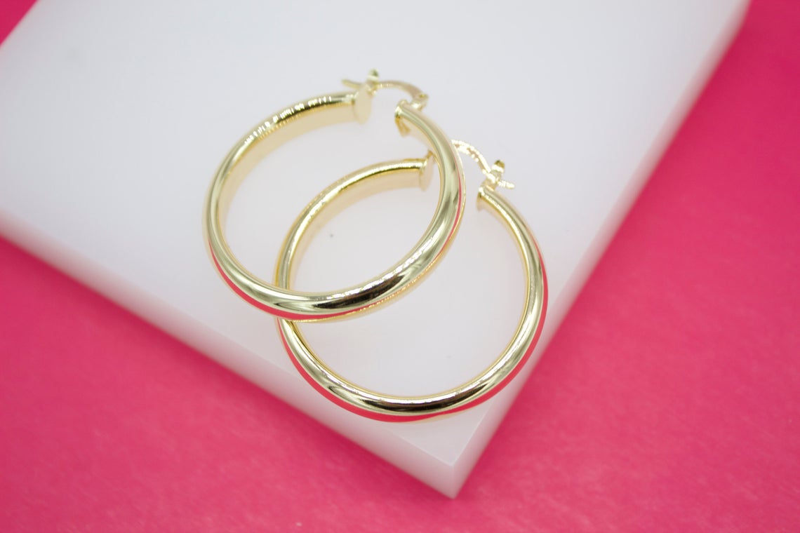 A pair of glossy gold hoop earrings rests neatly on a white display stand against a vibrant pink background.