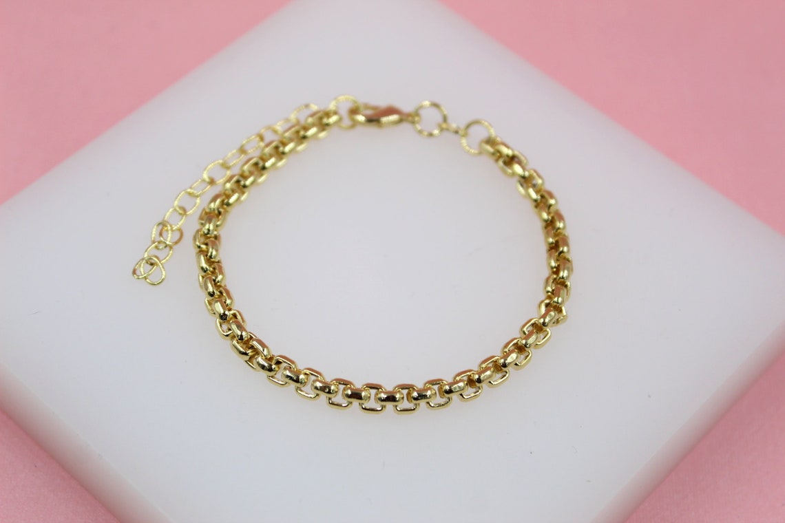 A delicate gold rolo chain bracelet rests on a white display block against a soft pink background.