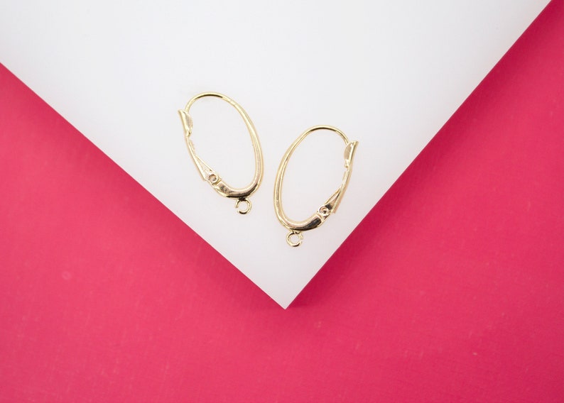 Two gold leverback earring findings with open loops rest on a white surface against a vibrant pink background.