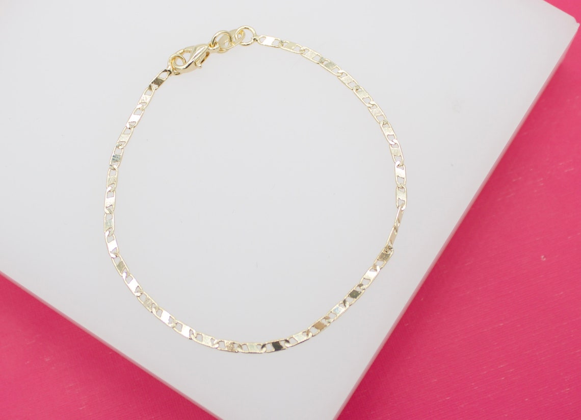 A delicate gold chain bracelet rests on a white display surface against a vibrant pink background.