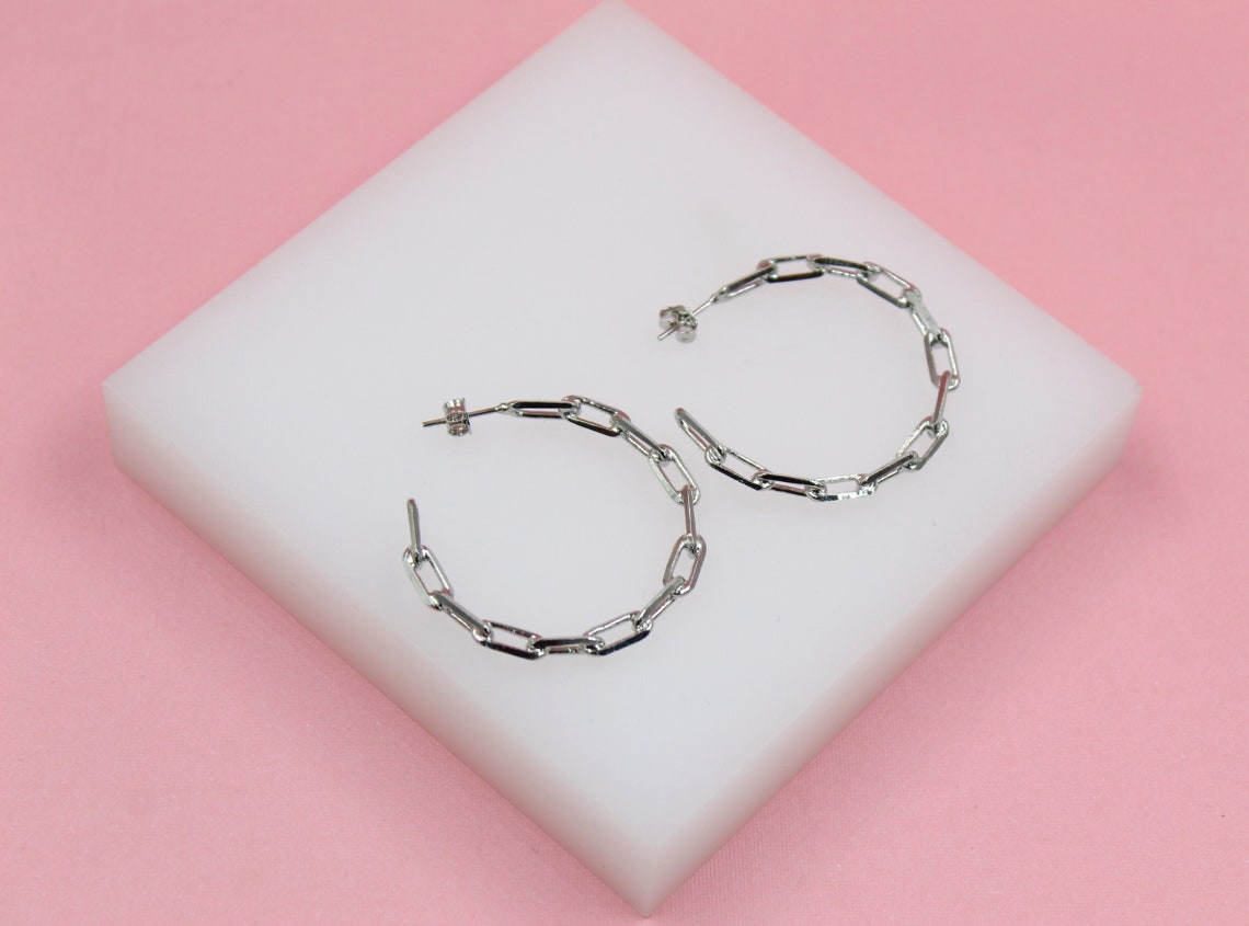 A pair of silver chain link hoop earrings rests on a white display block against a pink background.