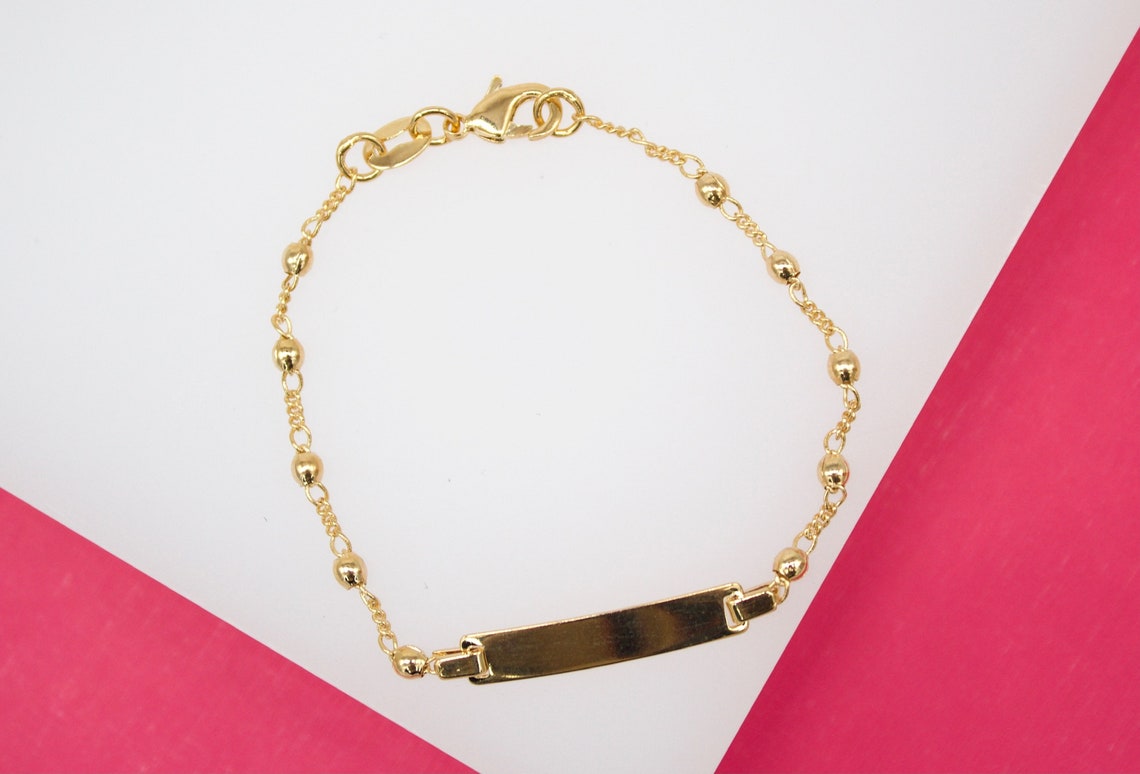 A delicate gold ID bracelet featuring small spherical beads and a blank rectangular plaque rests against a white and pink background.