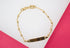 A delicate gold ID bracelet featuring small spherical beads and a blank rectangular plaque rests against a white and pink background.