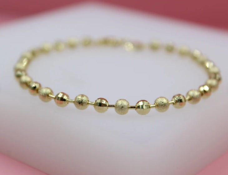 A delicate gold bead bracelet rests on a soft white surface against a blurred pink background.