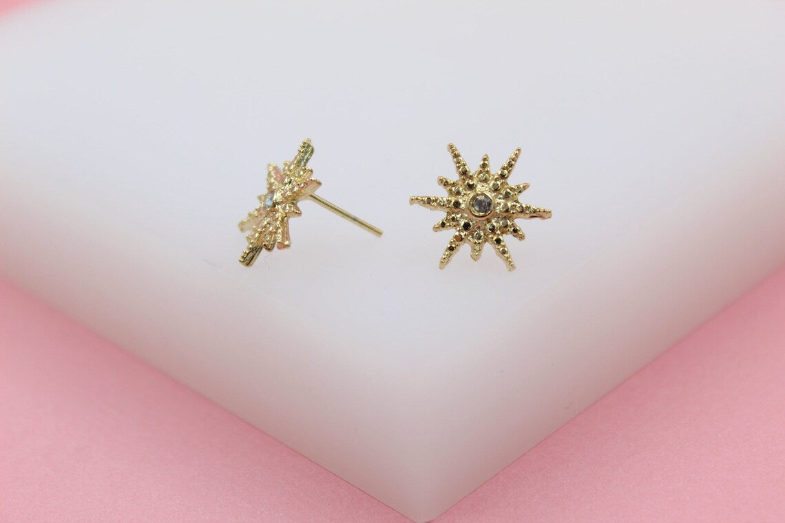 A pair of textured gold starburst stud earrings rests on a white display surface against a pink background.