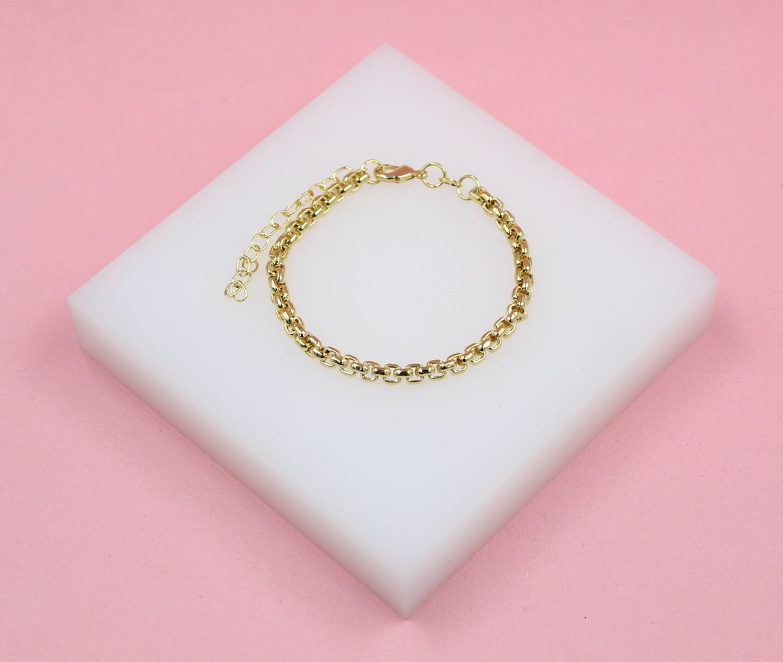 A dainty gold chain link bracelet rests on a square white display block against a soft pink background.