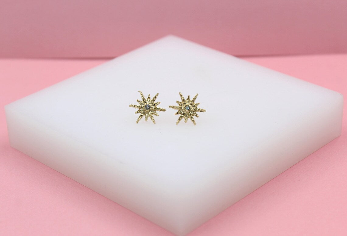 A pair of delicate gold starburst earrings with central blue gemstones rests on a white block against a pink background.
