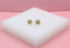 A pair of delicate gold starburst earrings with central blue gemstones rests on a white block against a pink background.
