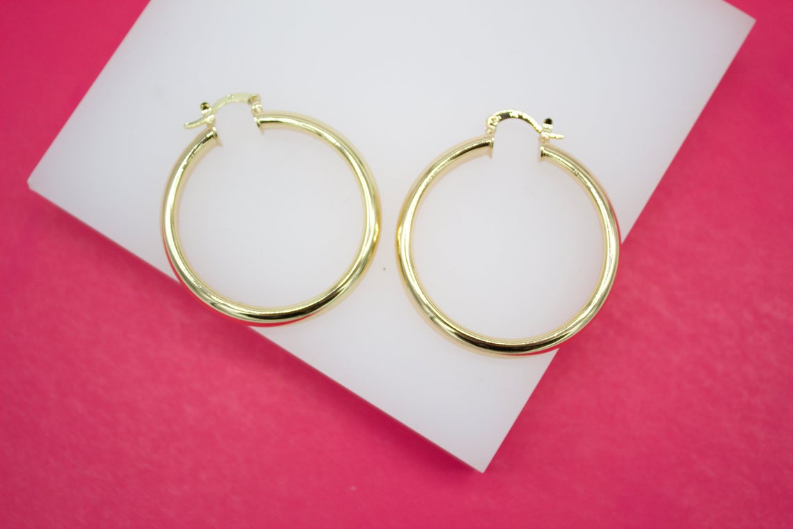 A pair of polished gold hoop earrings rests neatly on a white display stand against a vibrant pink background.