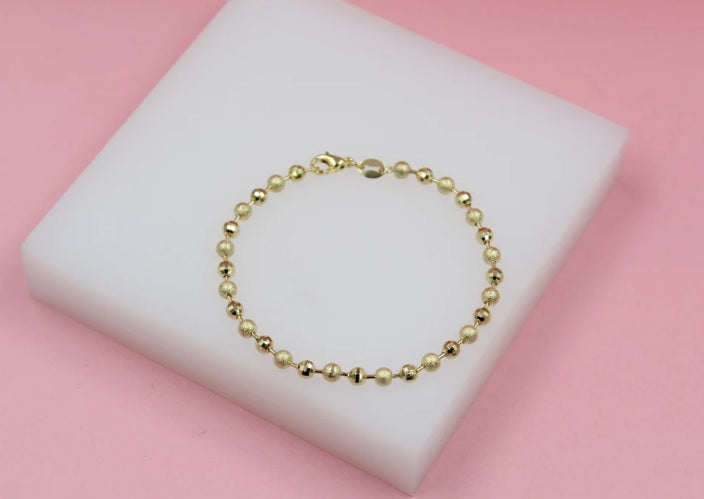 A delicate gold ball chain bracelet rests elegantly on a white display pad against a soft pink background.