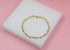 A delicate gold ball chain bracelet rests elegantly on a white display pad against a soft pink background.