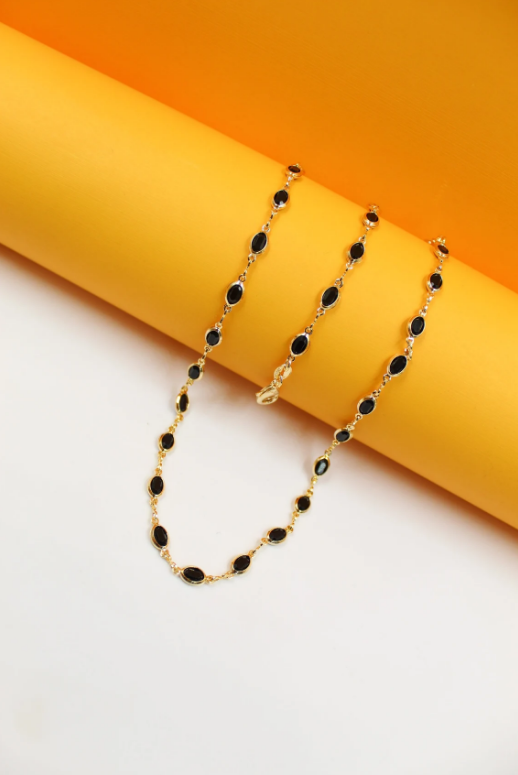 A delicate gold chain necklace featuring bezel-set black oval stones rests on a bright yellow and white geometric background.